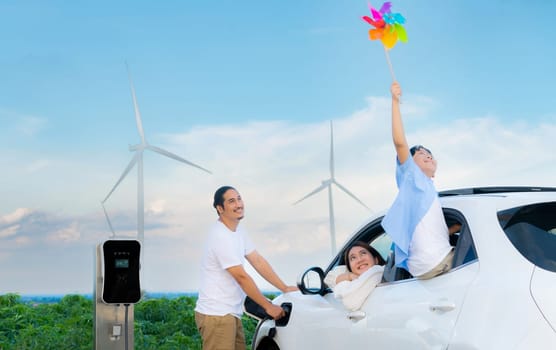 Concept of progressive happy family holding windmill toy and relax at wind farm with electric vehicle. Electric vehicle driven by clean renewable energy from wind turbine generator to charger station.