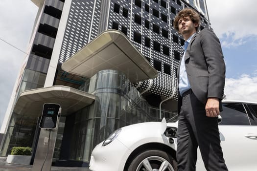 Below view of progressive businessman with electric car recharging at public charging station at modern city residential building. Eco friendly rechargeable EV car powered by alternative clean energy.