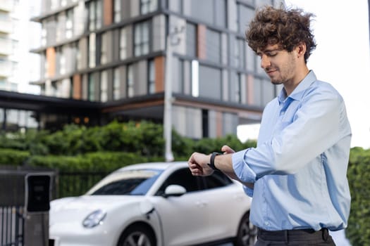 Businessman with smartwatch at modern charging station for electric vehicle with background of residential buildings as concept for progressive lifestyle of using eco-friendly as alternative energy.