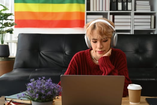 Young gay man wearing headphone and using laptop, communicating online, working remote from home.