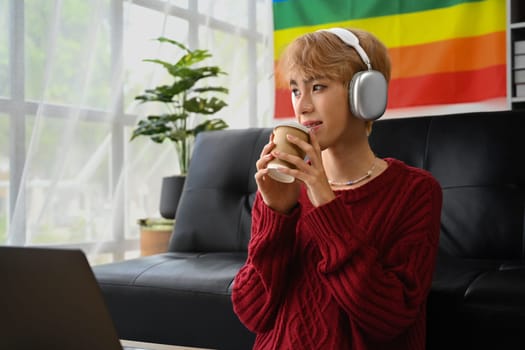 Peaceful young gay man wearing knitted sweater enjoy morning coffee and listening to music in wireless headphones.