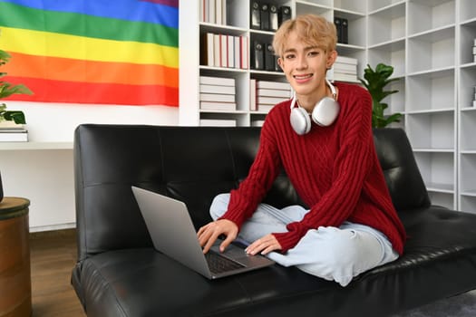 Young stylish asian gay man sitting on couch, working remotely, surfing internet on laptop in living room with rainbow flag on wall.