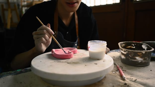 Cropped shot of man painting clay plate with a paintbrush at desk in ceramic workshop.