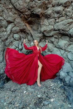 Red dress rocks woman. A blonde with flowing hair in a long flowing red dress stands near a rock of volcanic origin. Travel concept, photo session at sea.