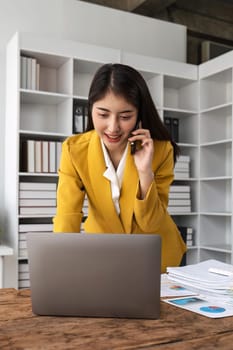 Portrait of young business woman with sitting in office in front of her laptop and talking on mobile phone.