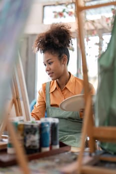 An African American woman who loves art or a female artist is holding a paint palette and paintbrushes while happily painting on paper at studio workshop.