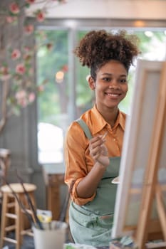 An African American woman who loves art or a female artist is holding a paint palette and paintbrushes while happily painting on paper at studio workshop.