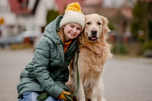 Preteen child girl hugging golden retriever dog at autumn city street wearing hat and warm jacket. Pretty kid with purebred pet doggy labrador outdoors