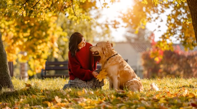 Beautiful girl with golden retriever dog sitting in autumn park with yellow leaves. Pretty young woman petting purebred doggy labrador at fall season at nature