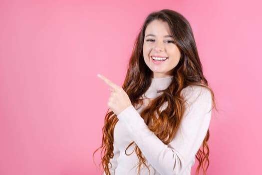Portrait of young happy woman smiling and standing isolated on pink background and pointing by finger on the empty space for advertising.