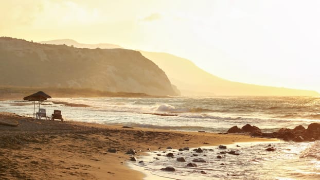 Two sunbeds on empty wild beach lit by golden sunset. Small waves over rock, hills in hazy background