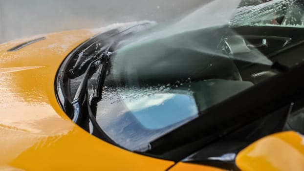 Yellow car windshield washed in self serve  carwash, water spraying from high pressure nozzle to front window.