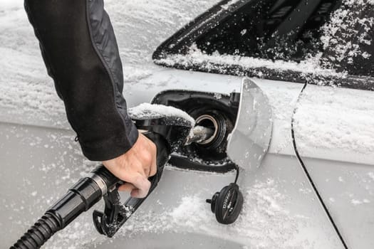 Detail on man's hand holding fuel nozzle, filling gas tank of car covered with snow in winter