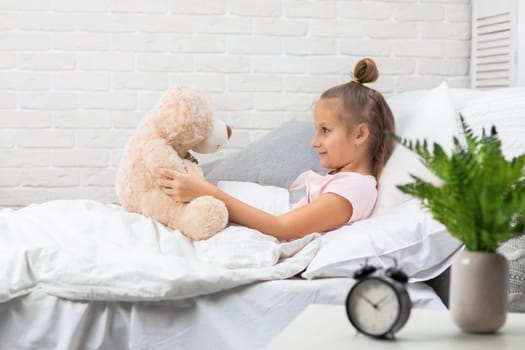 adorable little child girl playing with teddy bear in bed in morning