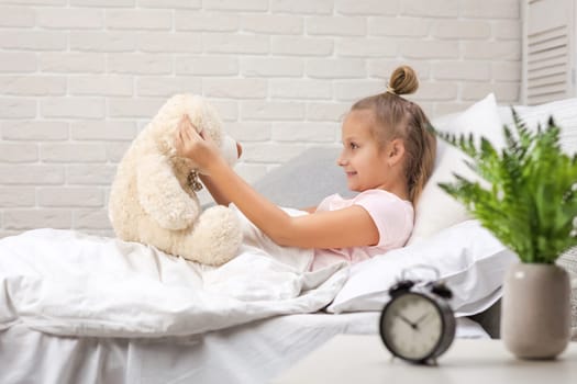 adorable little child girl playing with teddy bear in bed in morning