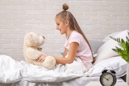 adorable happy little child girl playing with teddy bear in bed in morning.