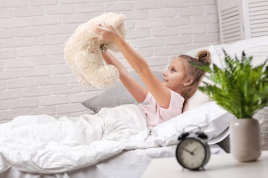 adorable little child girl playing with teddy bear in bed in morning