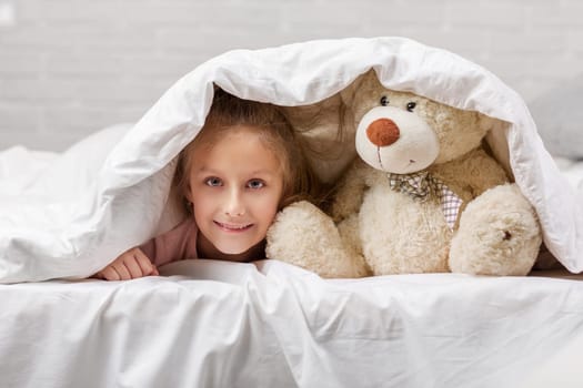 adorable little child girl playing with teddy bear in bed in morning. baby with a teddy bear under the covers