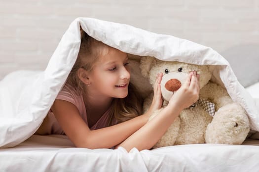 adorable little child girl playing with teddy bear in bed in morning. baby with a teddy bear under the covers