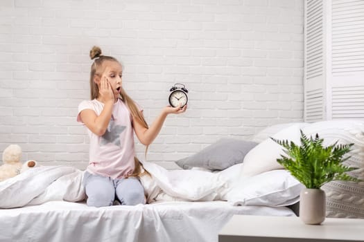 cute surprised little child girl in pyjamas with clock in bed. good morning