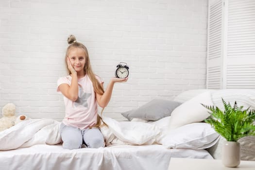 cute surprised little child girl in pyjamas with clock in bed. good morning