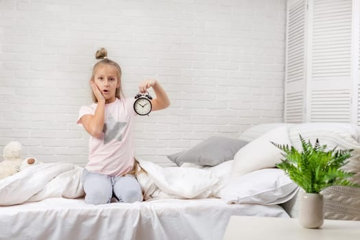 cute surprised little child girl in pyjamas with clock in bed. good morning