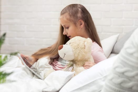 cute little child girl with teddy bear lies in bed uses digital tablet. child playing on tablet pc.