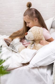 cute little child girl with teddy bear lies in bed uses digital tablet. child playing on tablet pc.