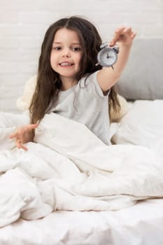 cute smiling little child girl in pyjamas with clock in bed. good morning