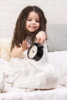 cute smiling little child girl in pyjamas with clock in bed. good morning