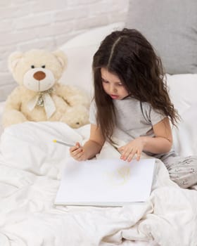 cute little girl with teddy bear drawing pictures while lying on bed. Kid painting at home