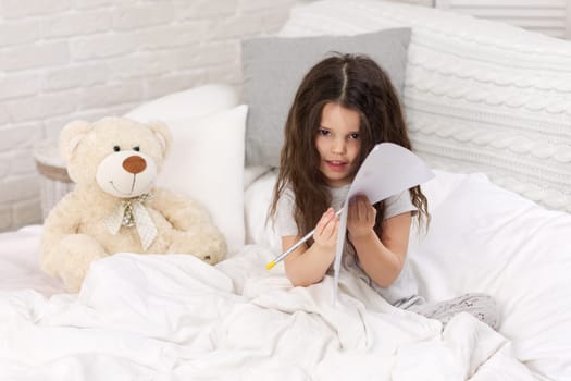 cute little girl with teddy bear drawing pictures while lying on bed. Kid painting at home