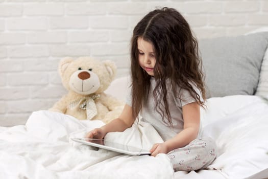 cute little child girl lies in bed uses digital tablet. child playing on tablet pc.