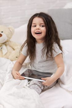 smiling little child girl lies in bed uses digital tablet. child playing on tablet pc.