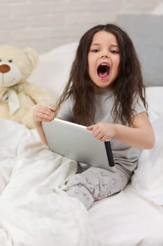 cute little child girl lies in bed uses digital tablet. child playing on tablet pc.