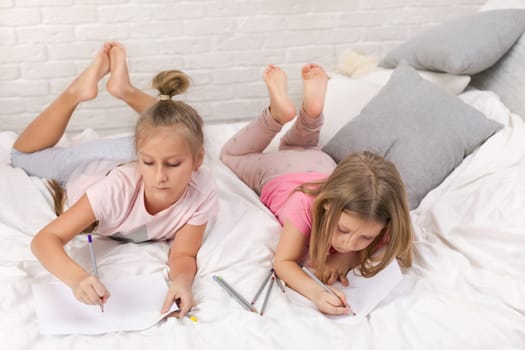 two cute children girls playing in the bedroom. kids drawing pictures while lying on bed. pajama party and friendship.