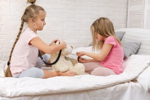 two cute children girls playing doctor with teddy bear at home. girl listens a stethoscope to toy.