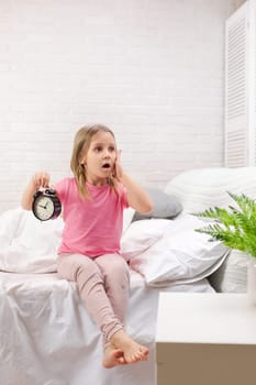 cute surprised little child girl in pyjamas with clock in bed. good morning