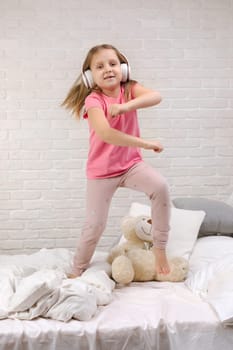 little child baby girl listening to the music with the headphones and dancing on bed. happy morning