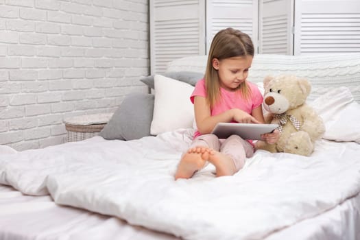 cute little child girl with teddy bear lies in bed uses digital tablet. child playing on tablet pc.