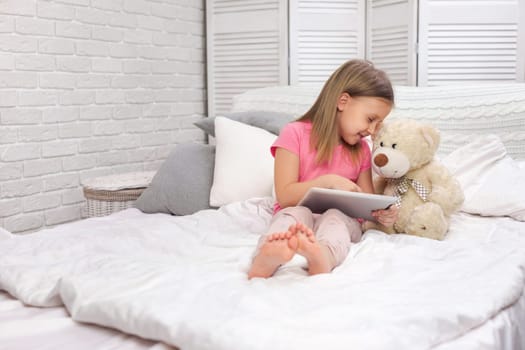 cute little child girl with teddy bear lies in bed uses digital tablet. child playing on tablet pc.
