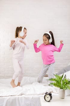 little children girls listening to the music with the headphones and dancing on bed. pajama party and friendship.