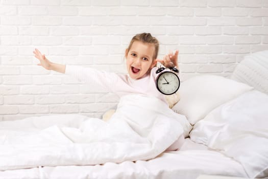 surprised little child girl in pyjamas with clock in bed. good morning