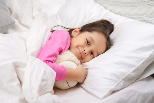 Cute happy little child girl sleeping with teddy bear in her bed
