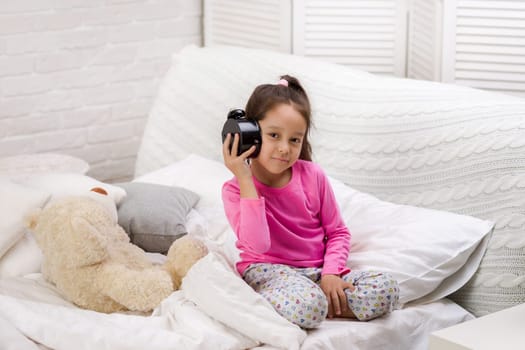 cute little child girl in pyjamas with clock in bed. good morning