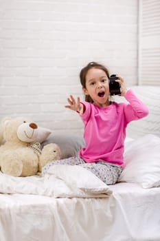 Surprised cute little child girl in pyjamas with clock in bed. good morning