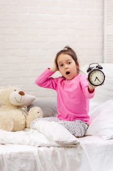 Surprised cute little child girl in pyjamas with clock in bed. good morning