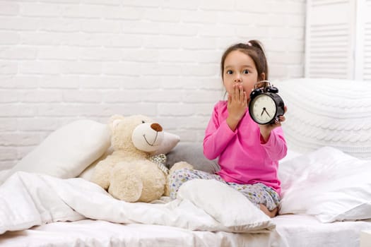 cute tired little child girl in pyjamas with clock in bed. good morning