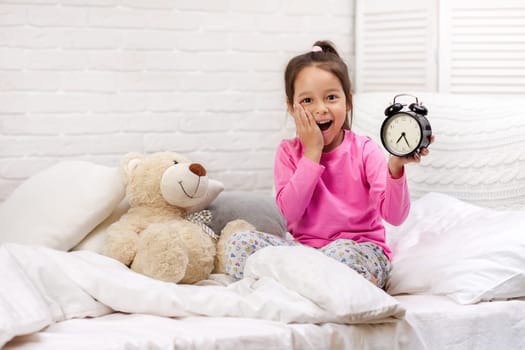 cute tired little child girl in pyjamas with clock in bed. good morning