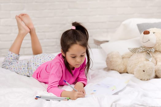 cute little girl drawing pictures while lying on bed. Kid painting at home